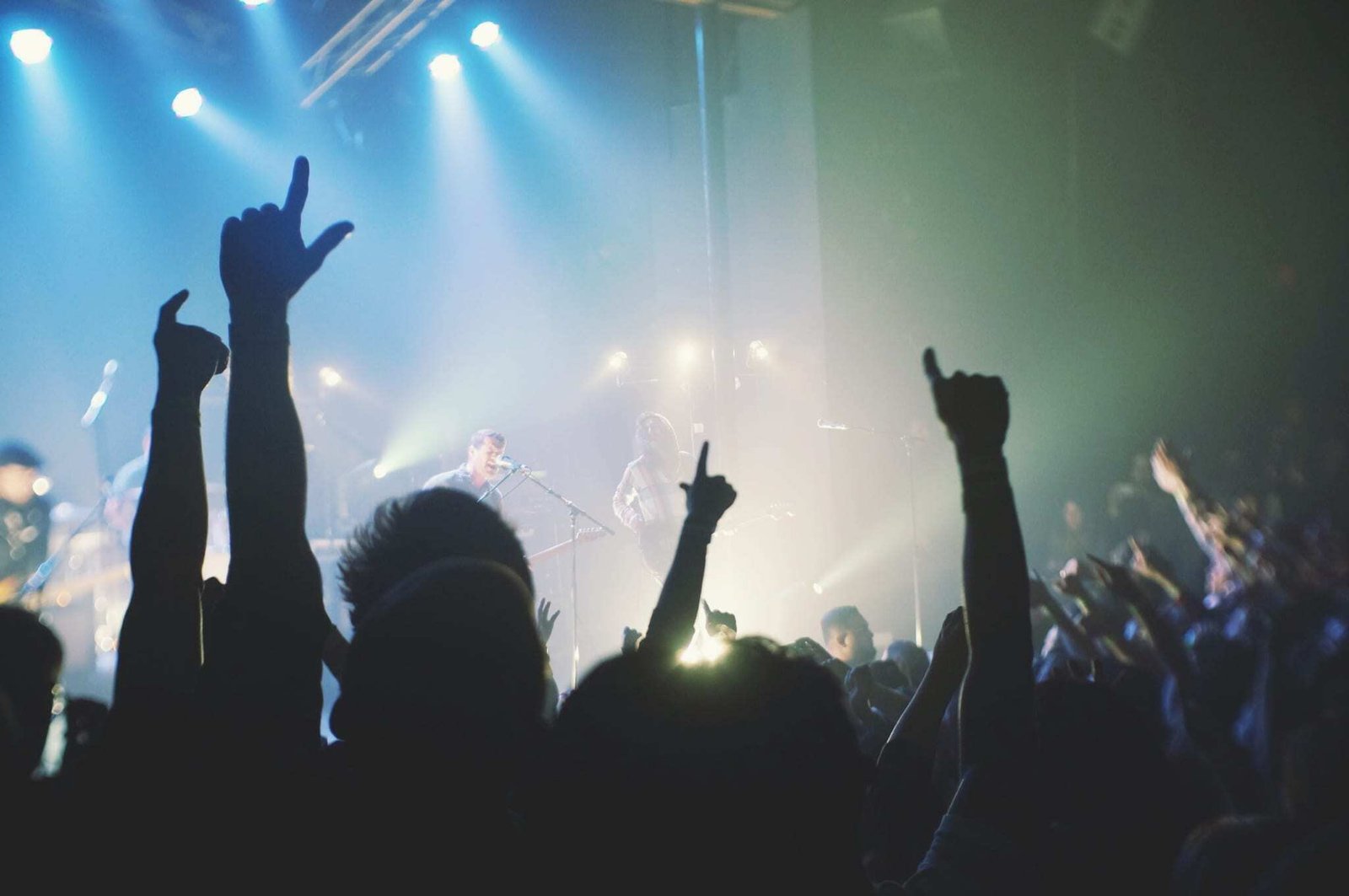 A crowd at a concert raises their hands in the air while a band performs on stage under blue and white lights, creating an energetic and lively atmosphere.