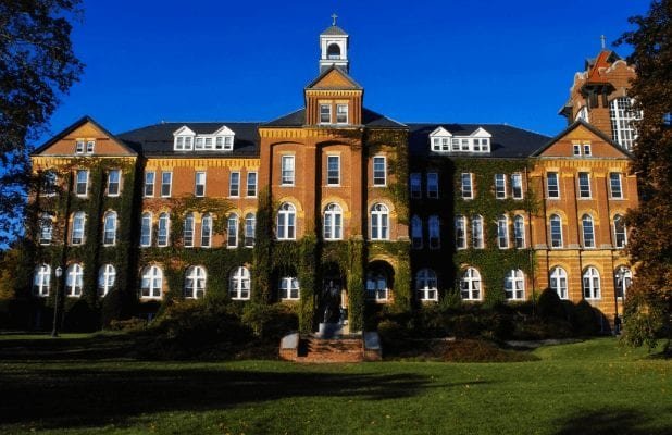 A large brick university building with ivy growing on its walls, tall arched windows, and a small central cupola, set against a clear blue sky and surrounded by green grass and trees.