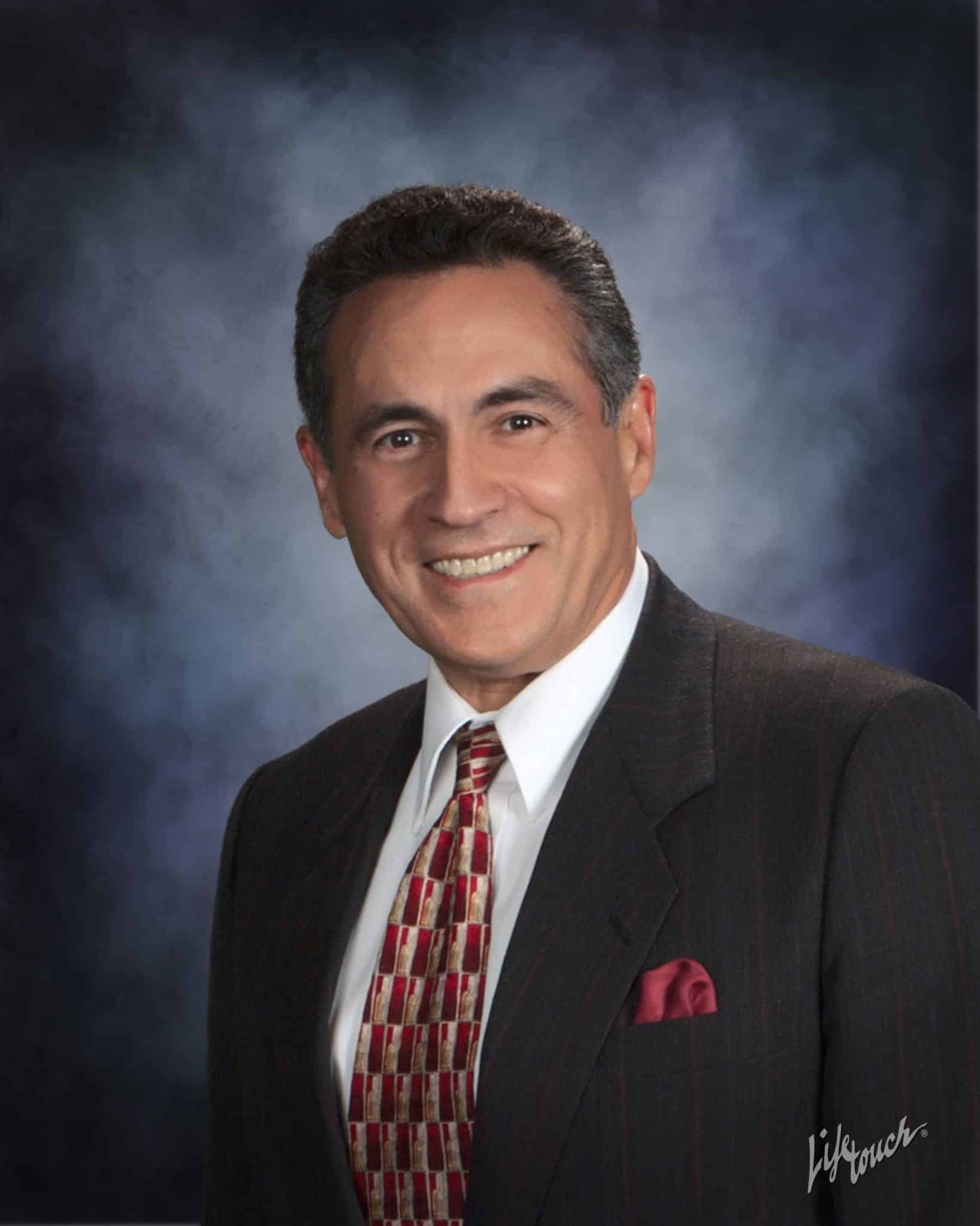 A man with dark hair wearing a dark suit, white shirt, and patterned red tie, smiles at the camera against a blue and purple gradient studio background.