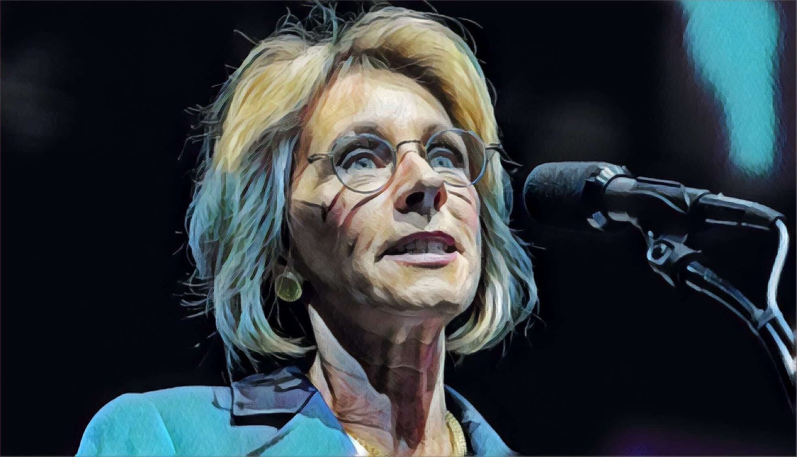 A woman with short blonde hair and glasses speaks into a microphone, wearing a blue blazer and gold earrings, against a dark background.
