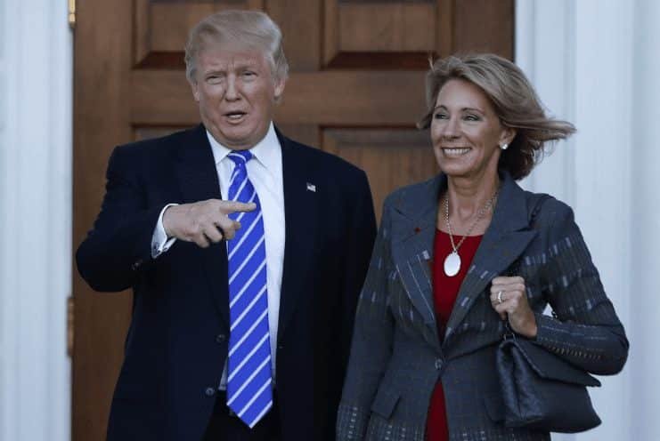 A man in a suit with a blue striped tie gestures while standing next to a woman in a gray blazer and red top. They are smiling in front of a large wooden door.