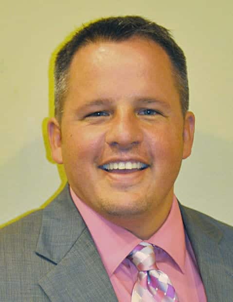 A smiling man wearing a gray suit jacket, pink dress shirt, and a pink patterned tie, posing in front of a plain light-colored background.