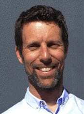 A man with short brown hair, a beard, and a mustache is smiling. He is wearing a light blue collared shirt and is standing in front of a plain gray background.