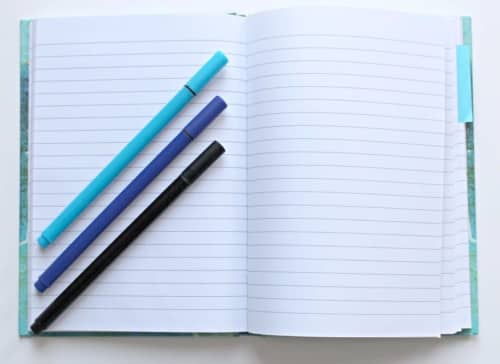 Open notebook with three pens laying diagonally at a 45 degree angle; blue, purple, black