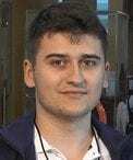 A young man with short dark hair is wearing a white shirt and a navy jacket. He is facing the camera and smiling slightly. The background is indoors and somewhat blurred.