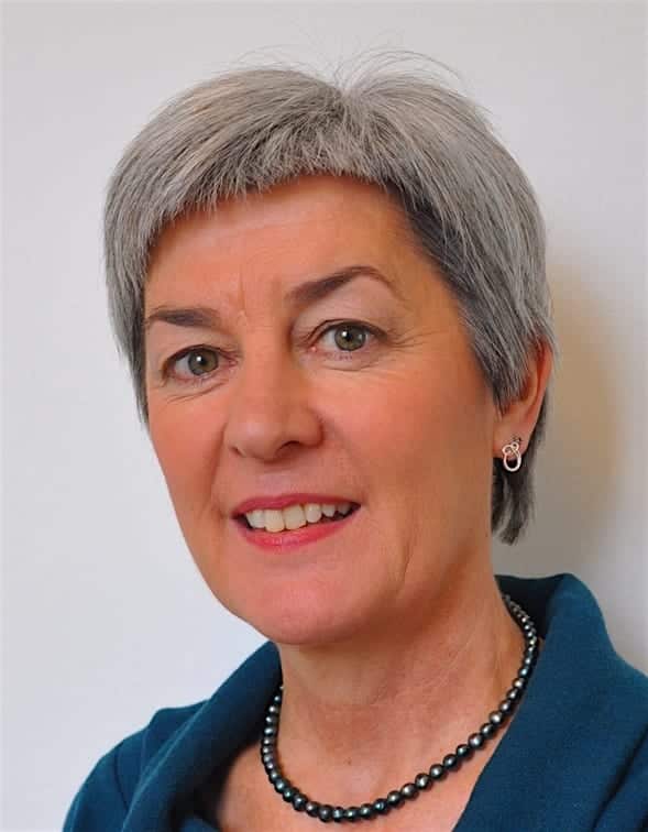 A woman with short gray hair, wearing a teal top, a pearl necklace, and small earrings, smiles in front of a plain white background.
