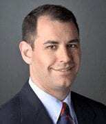 A man with short dark hair, wearing a dark suit jacket, white shirt, and a striped tie, is smiling at the camera against a plain gray background.