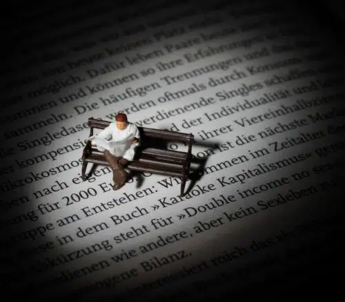 Tiny miniature of a man on a park bench on a page of a book reading a newspaper illuminated in a pool of light