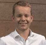 A young man with short blond hair, wearing a white button-up shirt, smiles at the camera while standing in front of a brown wooden wall.