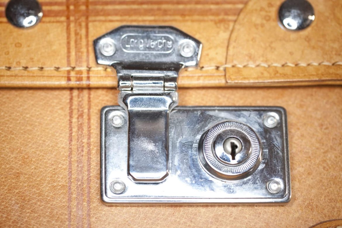 Close-up of a shiny silver latch and keyhole lock on a light brown leather suitcase or briefcase, showing stitching detail and metal hardware.