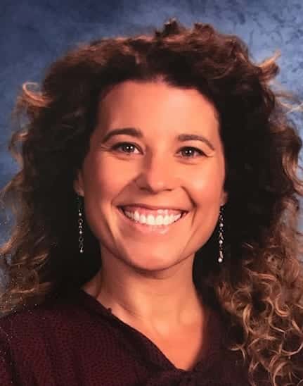 A woman with curly brown hair smiles at the camera. She is wearing a dark top and dangling earrings, with a blue textured background behind her.