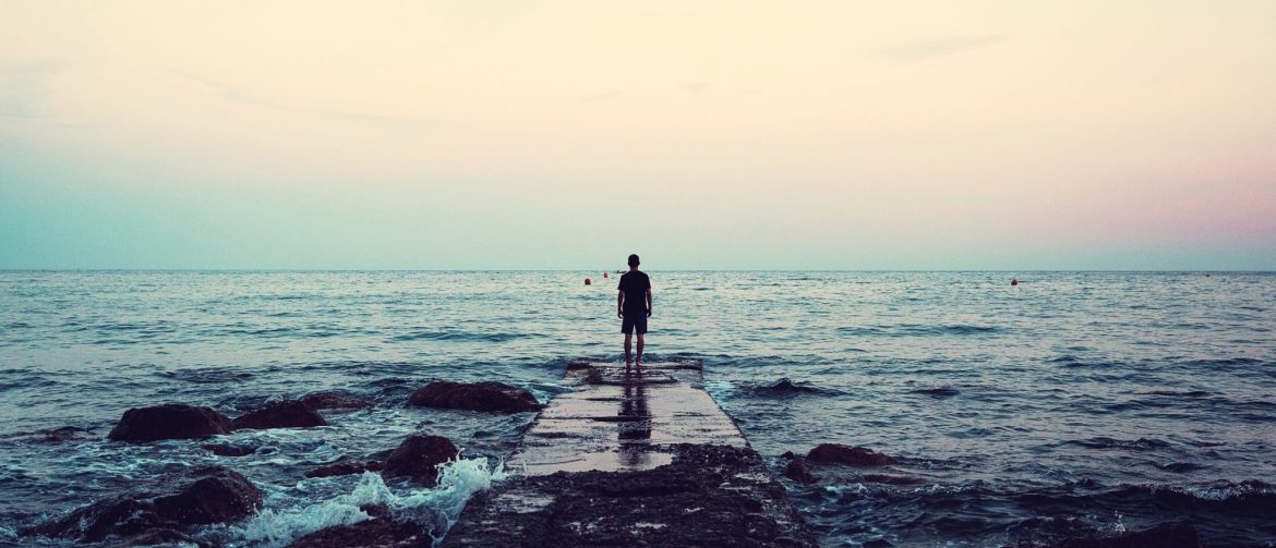 A person stands at the end of a rocky pier facing the calm sea under a pastel-colored sky at sunset, with gentle waves surrounding the rocks.
