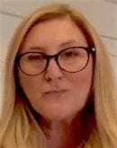 A woman with long blonde hair and black-rimmed glasses looks at the camera. She is indoors, and the background is softly lit and out of focus.