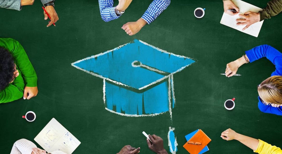Six people sit around a table with a large chalk drawing of a blue graduation cap in the center, along with notebooks, pens, and cups of coffee, suggesting a collaborative educational setting.