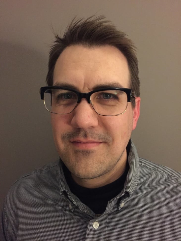 A man with short brown hair and glasses is smiling slightly. He is wearing a gray checkered button-up shirt over a black undershirt. The background is plain and light-colored.