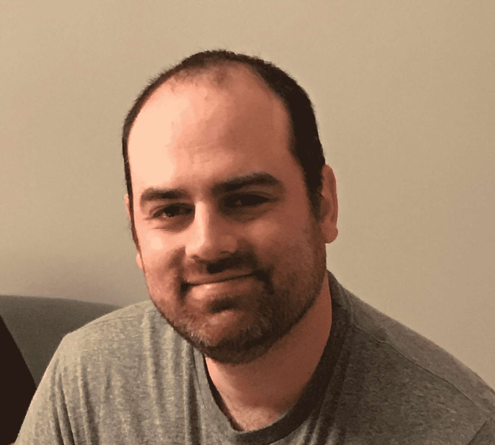 A man with short dark hair and a beard, wearing a gray t-shirt, smiles gently at the camera while seated indoors against a plain light-colored wall.