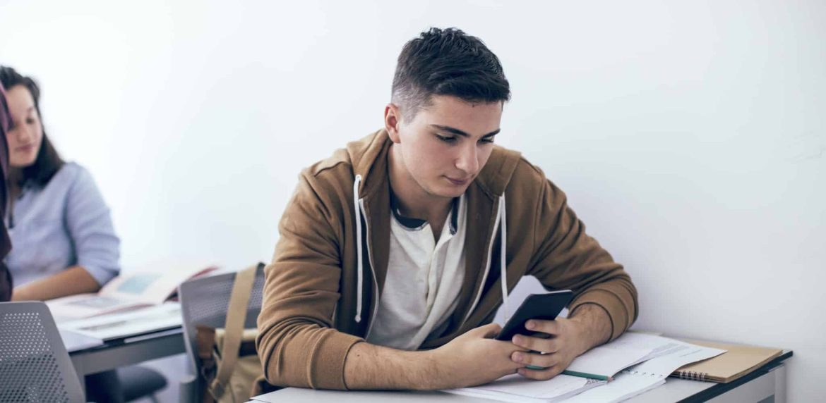 A student sitting at a desk in a classroom looks at a smartphone, with notebooks and papers spread out in front of him. Another student is seated at a nearby desk in the background.