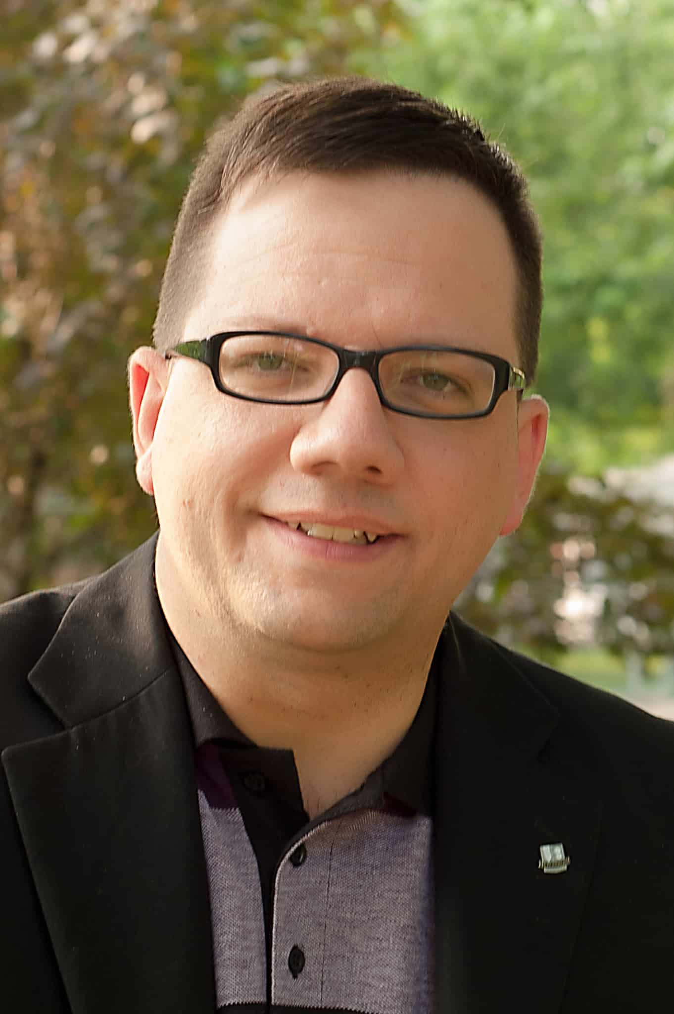 A man with short brown hair and glasses, wearing a dark blazer over a collared shirt, smiles at the camera outdoors with trees and greenery in the blurred background.