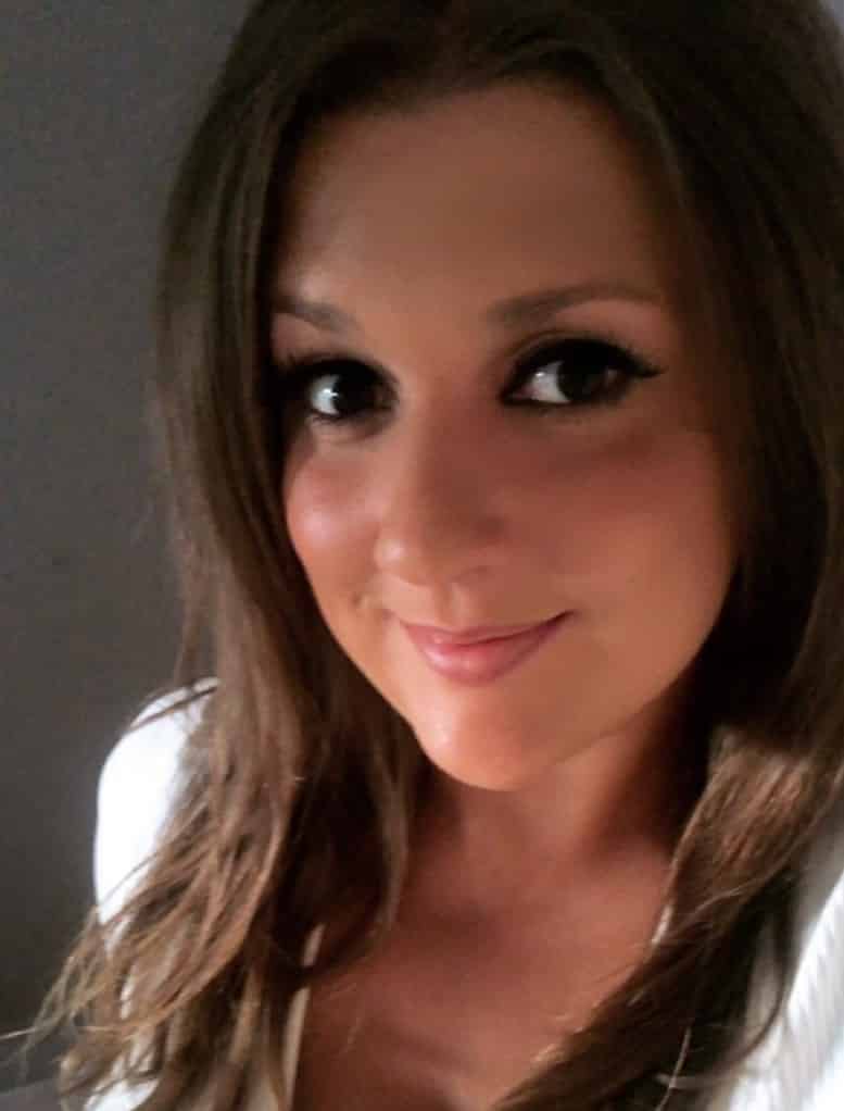 A woman with long brown hair and dark eyes smiles softly at the camera. She is wearing a white top and is posed against a neutral gray background.