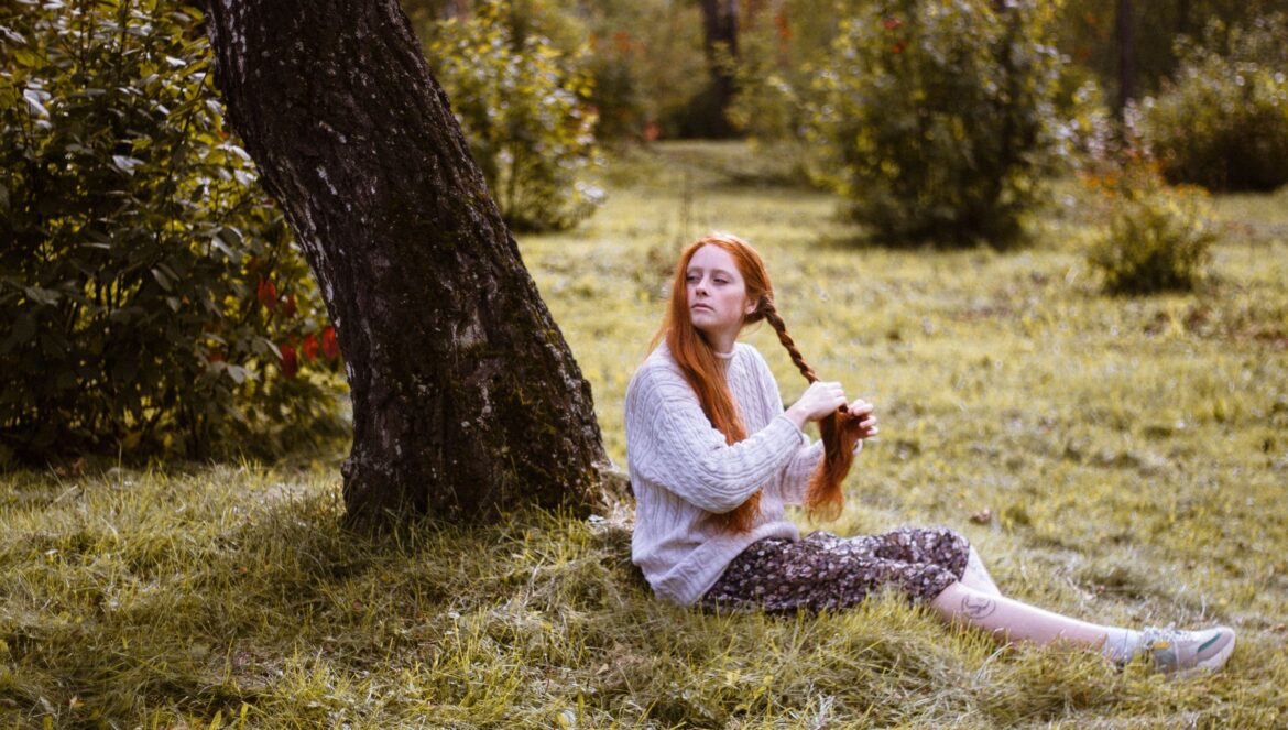 A young woman with red hair braiding her hair while sitting under a tree in a lush green park | edCircuit A young woman with red hair braiding her hair while sitting under a tree in a lush green park
