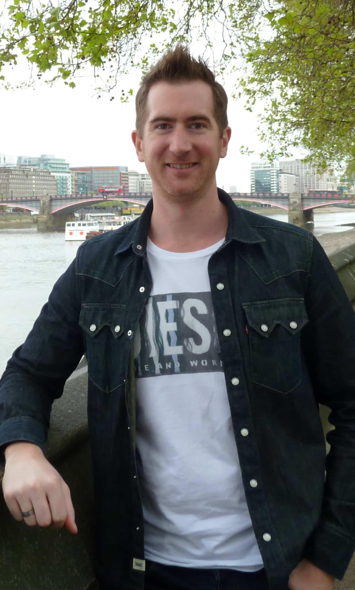 A man with short brown hair, wearing a black denim jacket over a white graphic t-shirt, stands and smiles by a riverside with a cityscape and a bridge in the background.