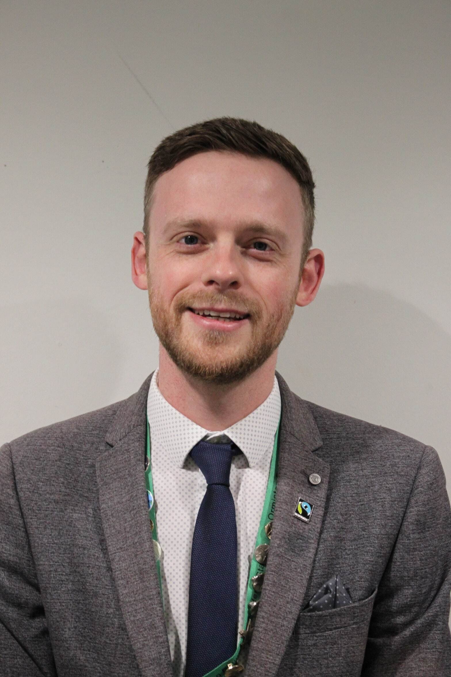 A man with short brown hair and a trimmed beard, wearing a grey suit jacket, white shirt, navy tie, and green lanyard, smiles at the camera in front of a plain light background.