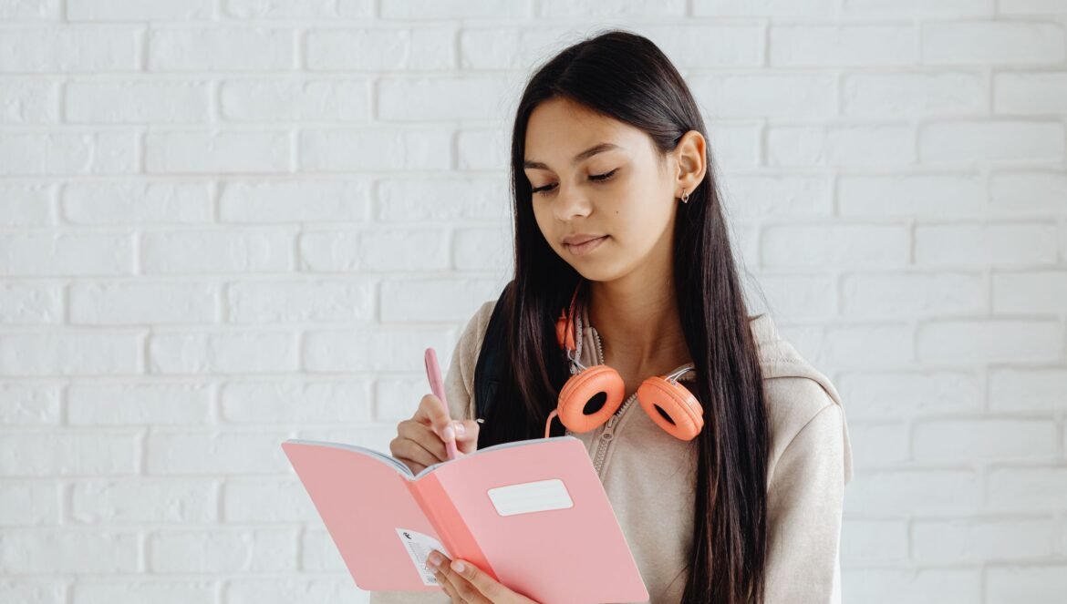 Teen girl writing notes in a pink notebook with headphones Indoors setting | edCircuit Teen girl writing notes in a pink notebook with headphones Indoors setting