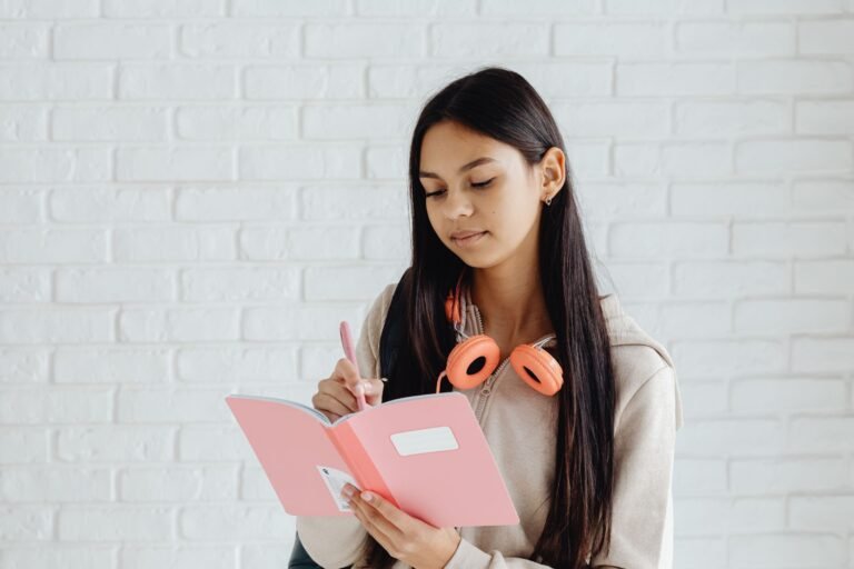 Teen girl writing notes in a pink notebook with headphones. Indoors setting