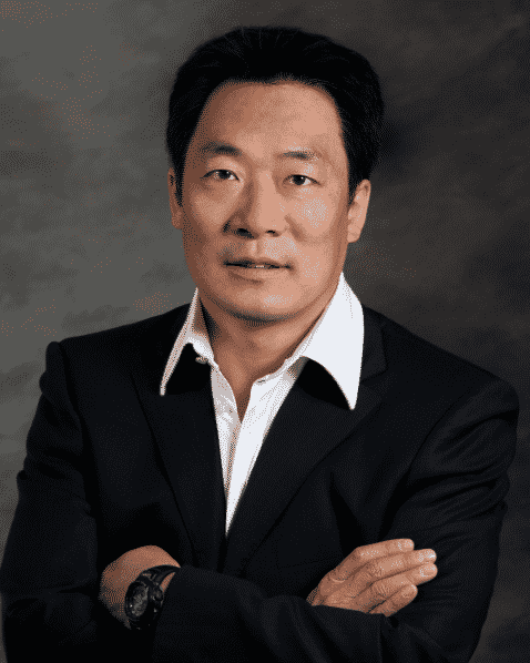 A man in a black suit with a white dress shirt stands with arms crossed, looking at the camera against a neutral, dark background.