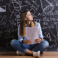 A young woman sits cross-legged on the floor, holding notes and looking thoughtfully at a blackboard covered in complex math and science equations and diagrams.