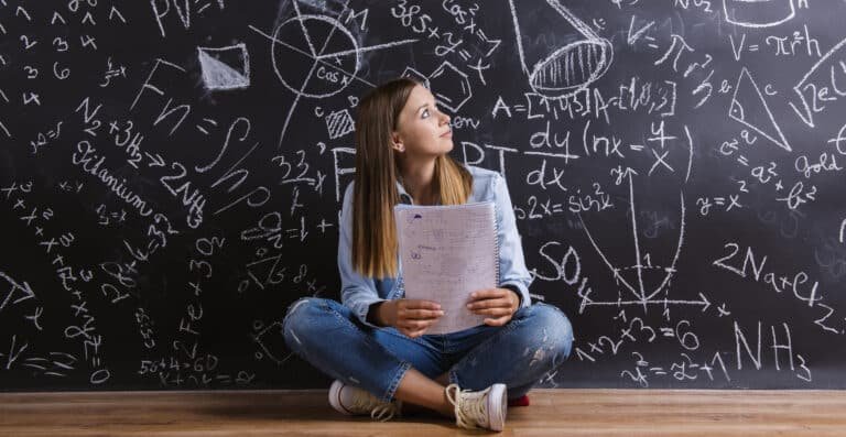 A young woman sits crosslegged on the floor holding notes and looking thoughtfully at a blackboard covered in complex math and science equations and diagrams | edCircuit A young woman sits cross legged on the floor holding notes and looking thoughtfully at a blackboard covered in complex math and science equations and diagrams