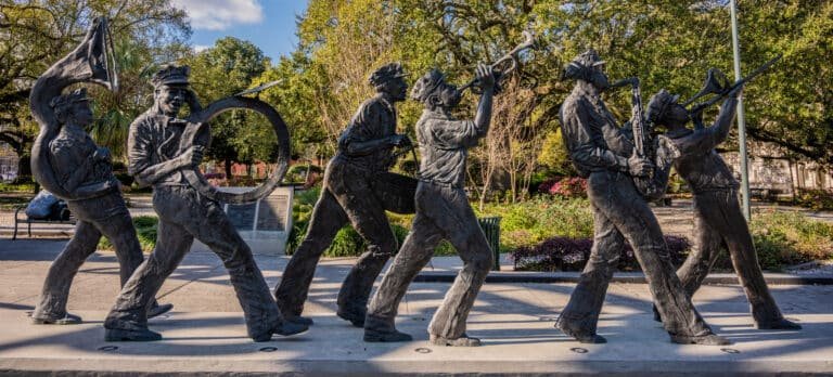 A bronze sculpture of six jazz musicians in a lively parade formation, each playing instruments like trumpet, trombone, saxophone, drum, and tuba, set outdoors in a park with trees and flowers in the background.