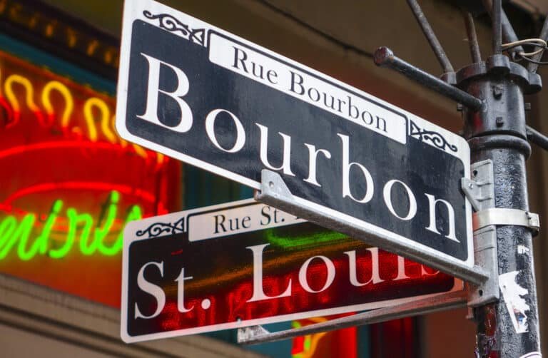 Close-up of intersecting French-inspired street signs for Bourbon Street and St. Louis Street, with colorful neon lights blurred in the background.