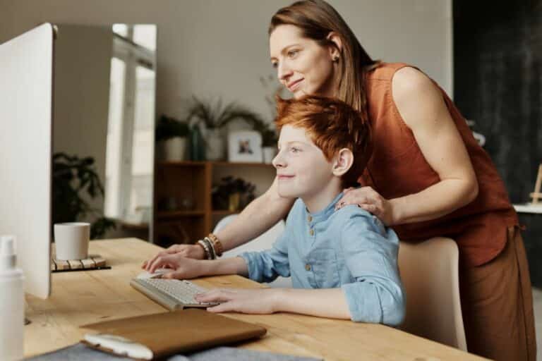 A woman stands behind a smiling boy who is seated at a desk, using a computer. The woman rests her hands on his shoulders, and they both look at the computer screen in a bright, cozy room.