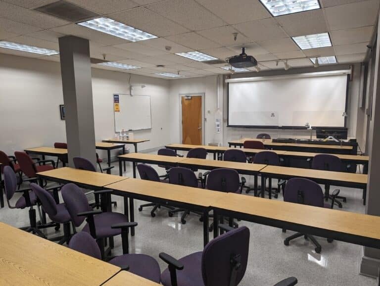 A classroom with rows of empty wooden desks and purple chairs, a whiteboard and projector screen at the front, and fluorescent lights on the ceiling. Bottles and supplies are on a table near the door.