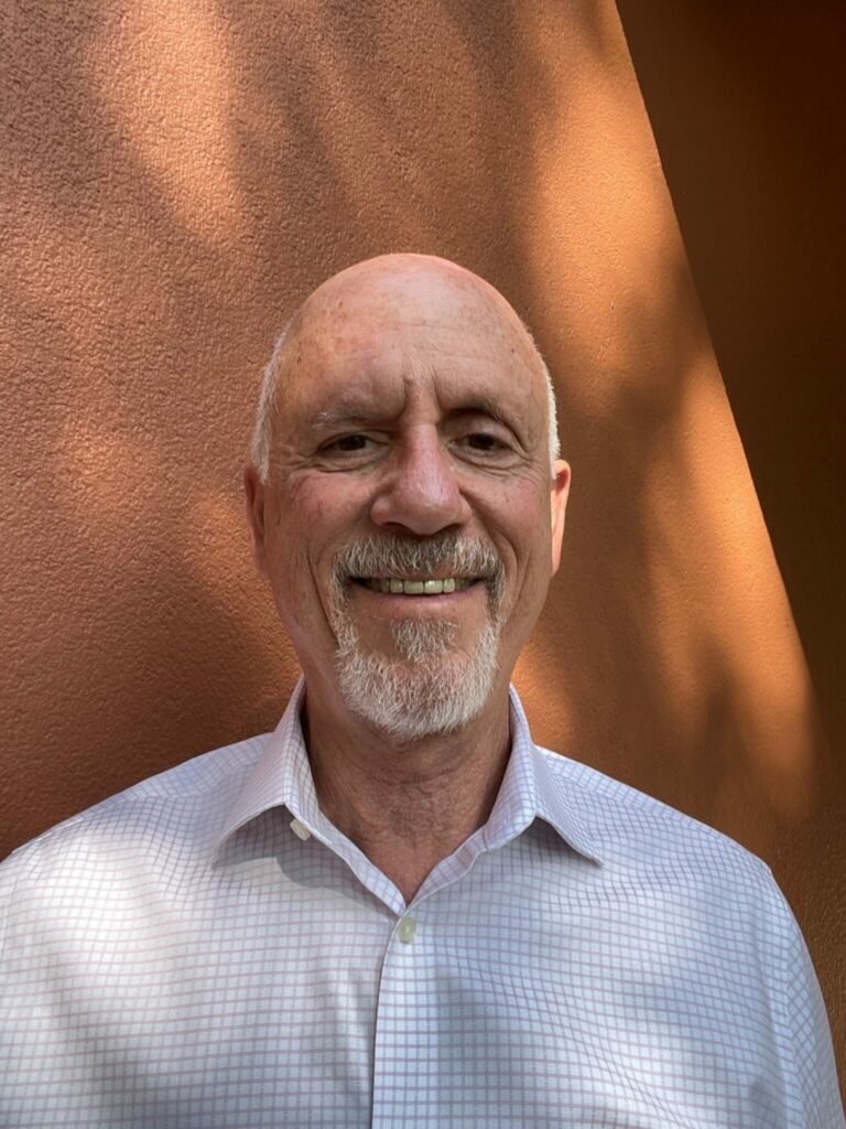 An older man with a bald head and white beard smiles at the camera. He is wearing a light-colored, checkered dress shirt and stands in front of a sunlit, peach-colored textured wall.