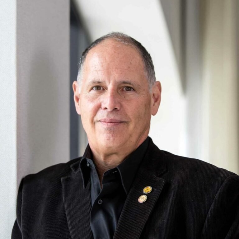 A middle-aged man with short gray hair wearing a dark blazer and black shirt stands indoors, smiling slightly, with a neutral background and soft lighting.