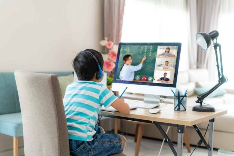 A young boy wearing headphones sits at a desk, looking at a computer screen showing an online class with a teacher writing on a chalkboard and several students visible in video call windows.