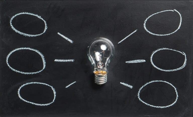 A light bulb on a blackboard is surrounded by six empty ovals drawn in chalk, connected to the bulb with lines, representing ideas or brainstorming.