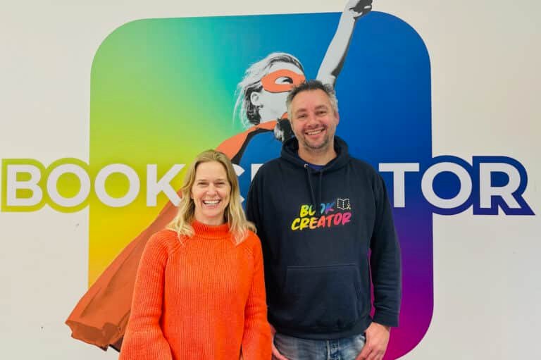 Two people smiling in front of a colorful wall with the word BOOK CREATOR and an image of a superhero figure in a red cape and mask. The woman wears an orange sweater; the man wears a dark hoodie with Creator on it.