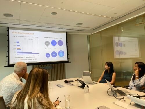 Four people sit around a conference table in a meeting room, looking at a large screen displaying presentation slides about Southeast Asia’s economy and internet growth. Laptops and papers are on the table.