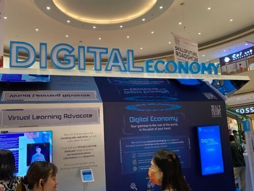 People stand in front of a DIGITAL ECONOMY exhibit at a mall, featuring informational panels about virtual learning and digital initiatives. Bright blue signage and interactive displays are visible.