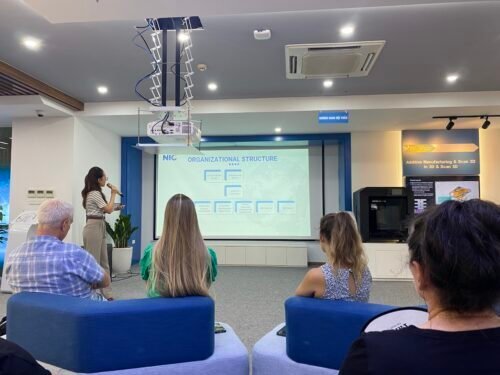 A woman stands at the front of a modern room giving a presentation on organizational structure, as four people seated in chairs listen and look at the large screen displaying a flowchart.