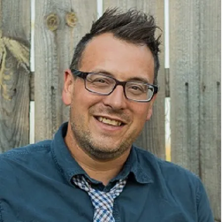 A man with short, dark, spiked hair, glasses, and a blue shirt with a checkered tie smiles in front of a wooden fence.