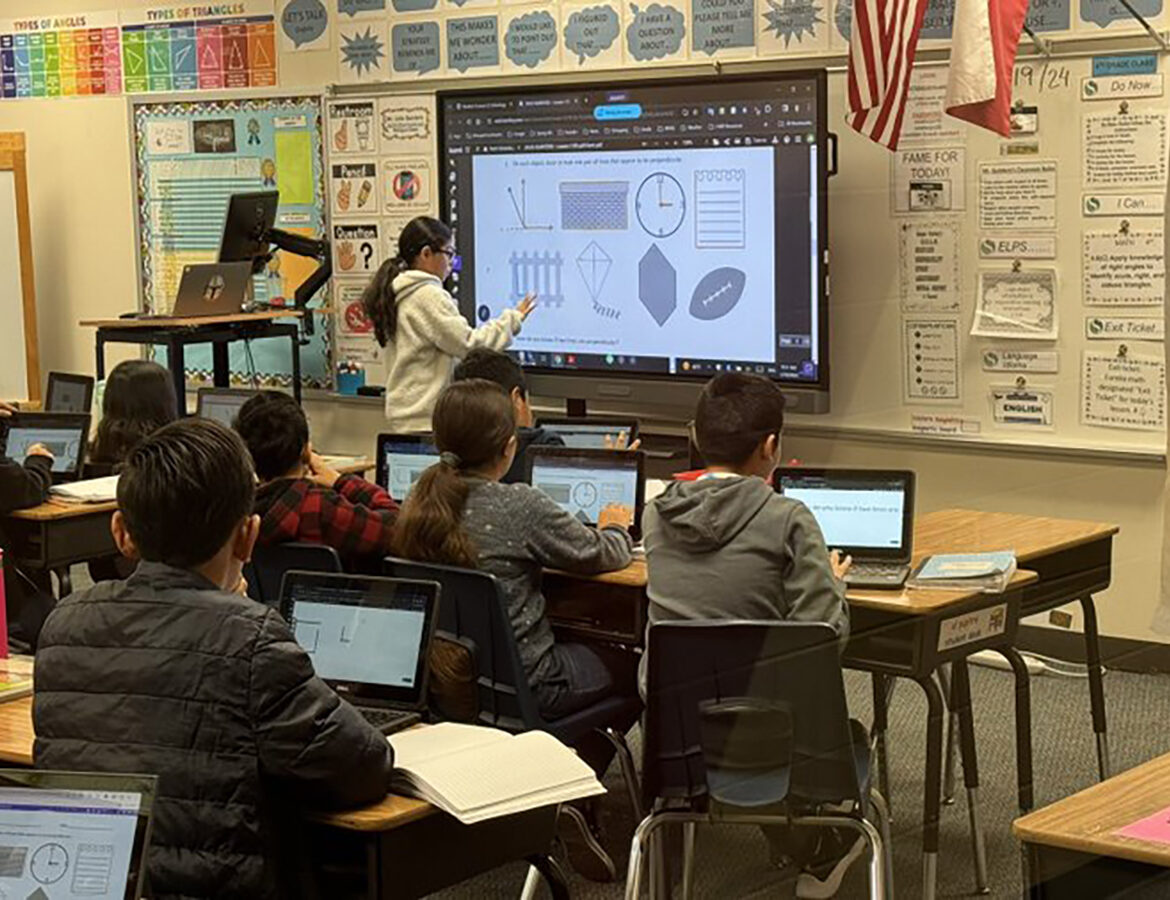 A teacher uses a digital whiteboard to explain math concepts to a classroom of students, who are seated at desks working on laptops and paying attention to the lesson.