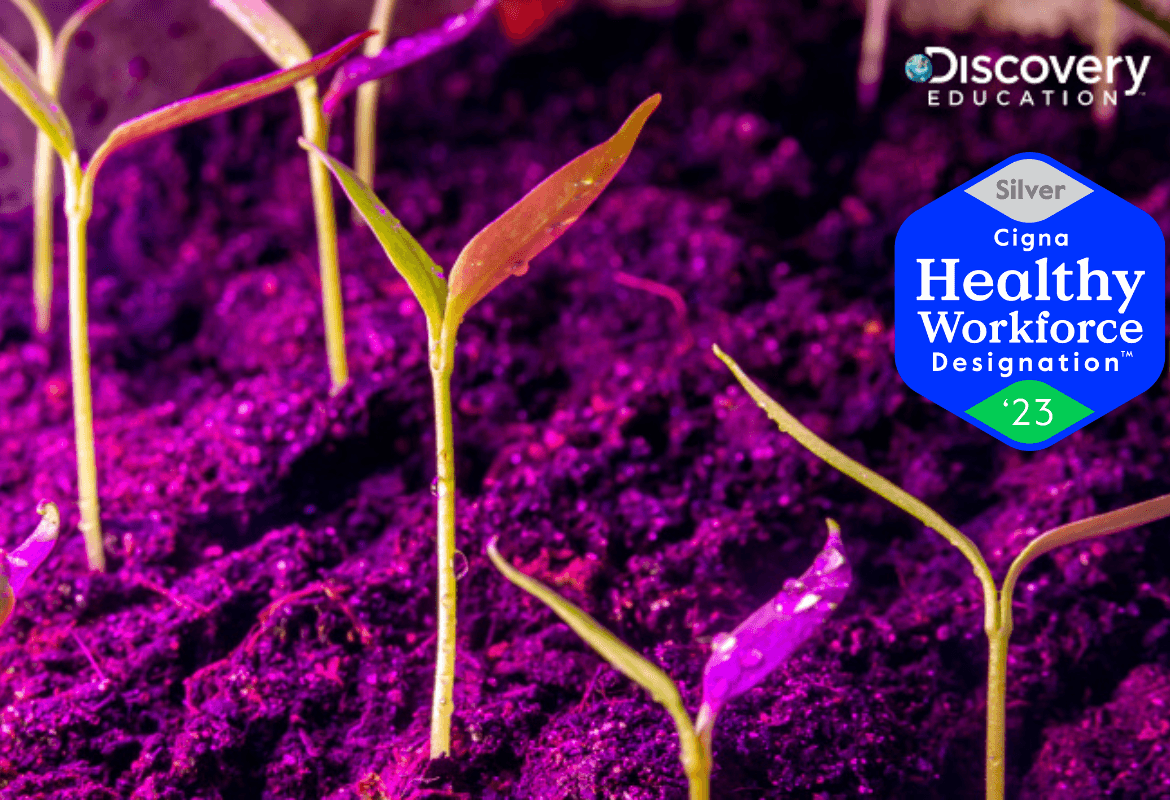 Young green seedlings sprout from moist, purple-lit soil. The Discovery Education logo appears in the top right, along with a silver “Cigna Healthy Workforce Designation 2023” badge.