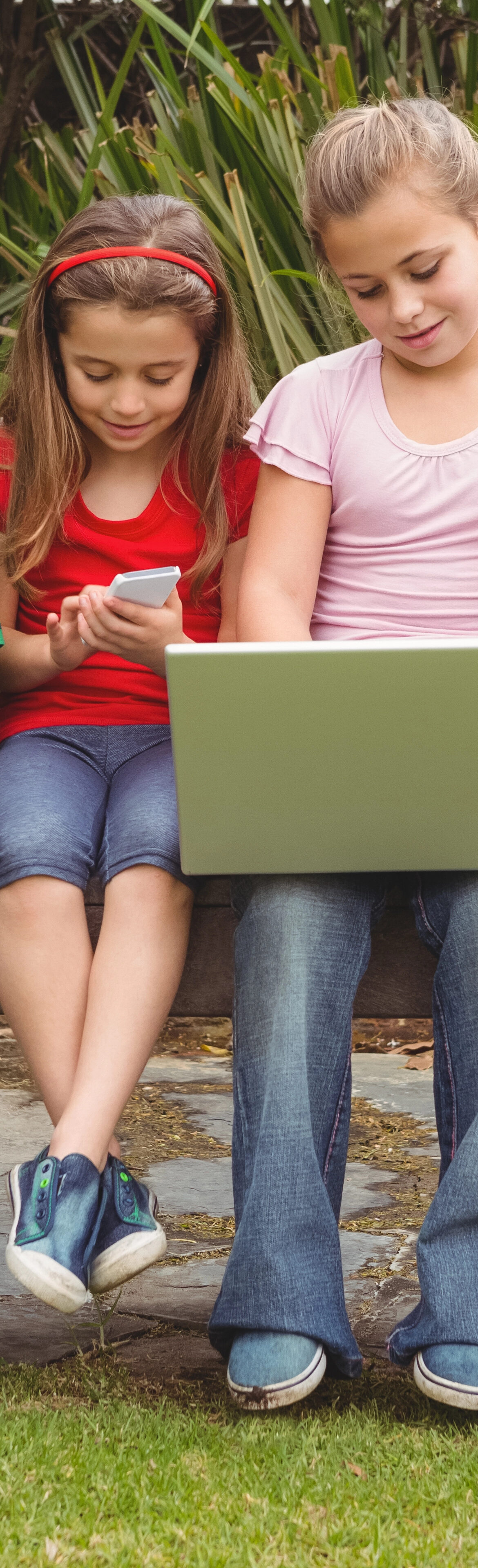 Six children sitting on a wooden bench outside, using various electronic devices such as laptops and tablets, with greenery in the background. They appear focused and engaged.