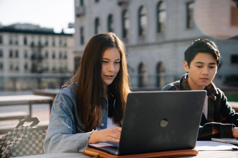 Two students collaborating on a laptop outdoors with a historic buildi