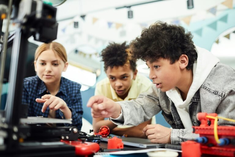 Three children closely observe and adjust a 3D printer, surrounded by various tools and colorful parts, in a bright classroom decorated with hanging flags.