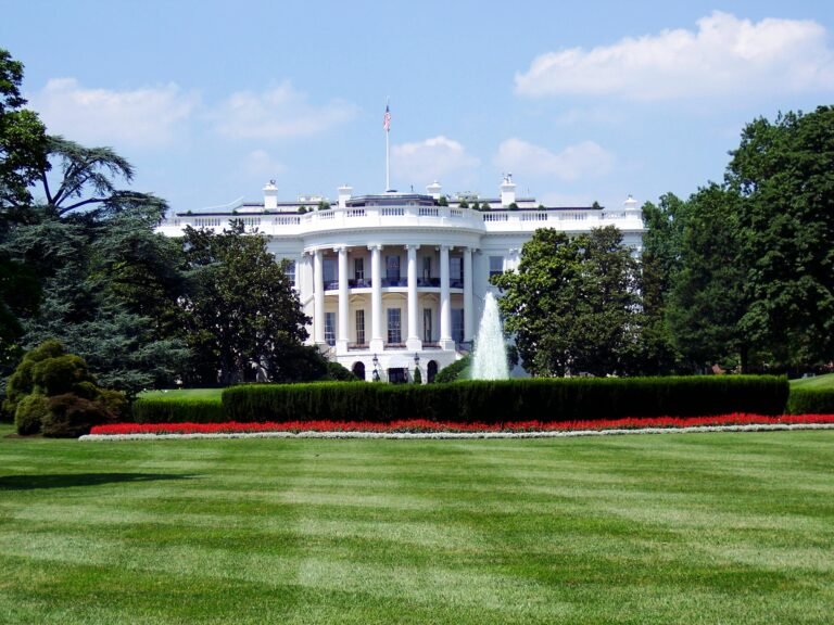 The White House stands behind a lush green lawn, trimmed hedges, and colorful flowerbeds. A fountain is in front, with several trees and a clear, partly cloudy sky in the background.
